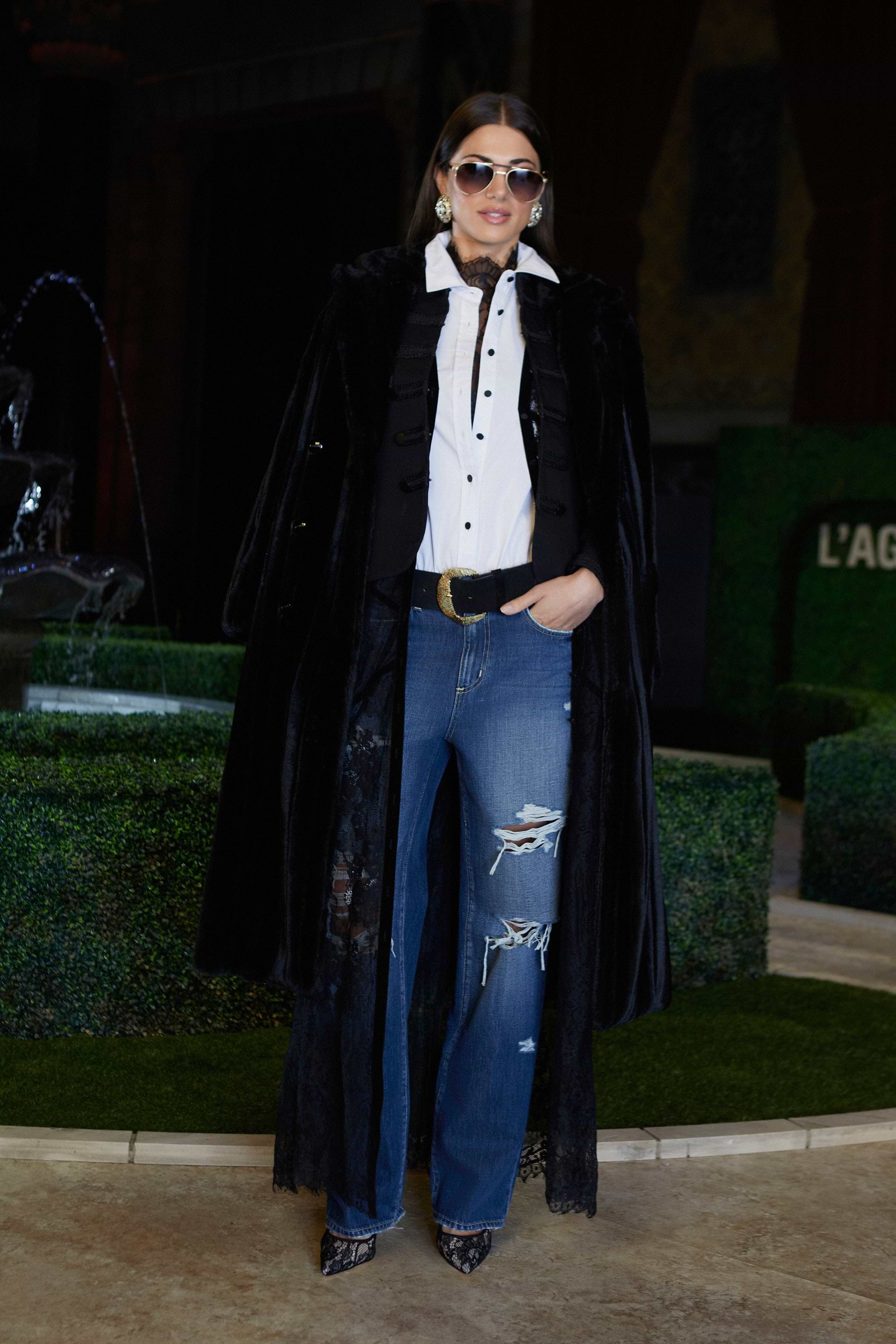 A woman wearing sunglasses, a white button-up shirt, a black long coat, blue ripped jeans, and heels stands confidently indoors near a decorative green hedge and a fountain.