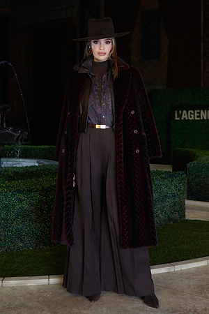 A woman stands outdoors wearing a dark brown wide-brim hat, a long textured burgundy coat, a paisley-patterned blouse, high-waisted brown pleated trousers, and brown shoes. Green decorative hedges and a fountain are behind her.