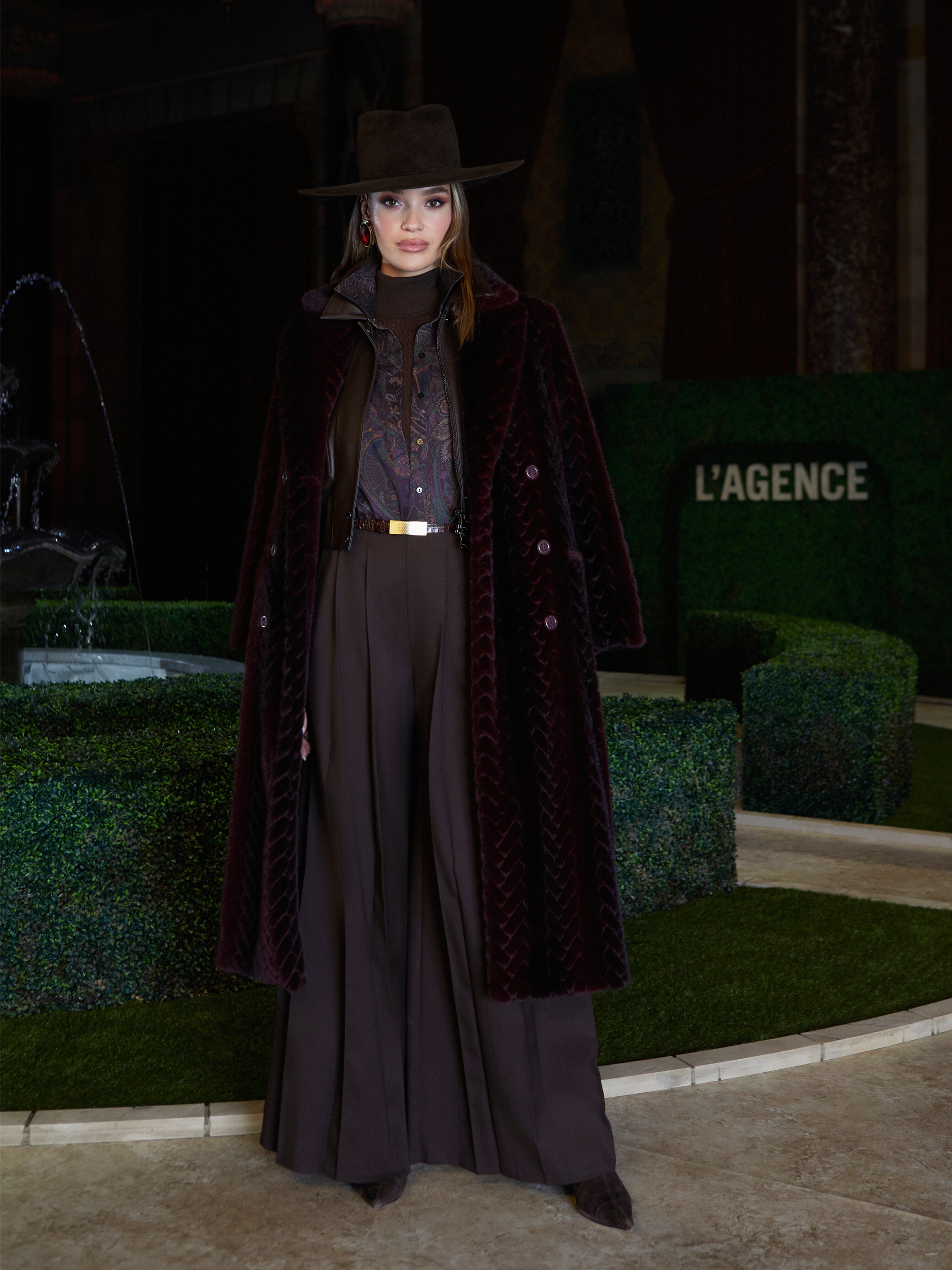 A woman in the Icelyn Long Coat channels historical romance by a fountain and green hedges with LAGENCE behind her, evoking Fall 2026 New York Fashion Week elegance.