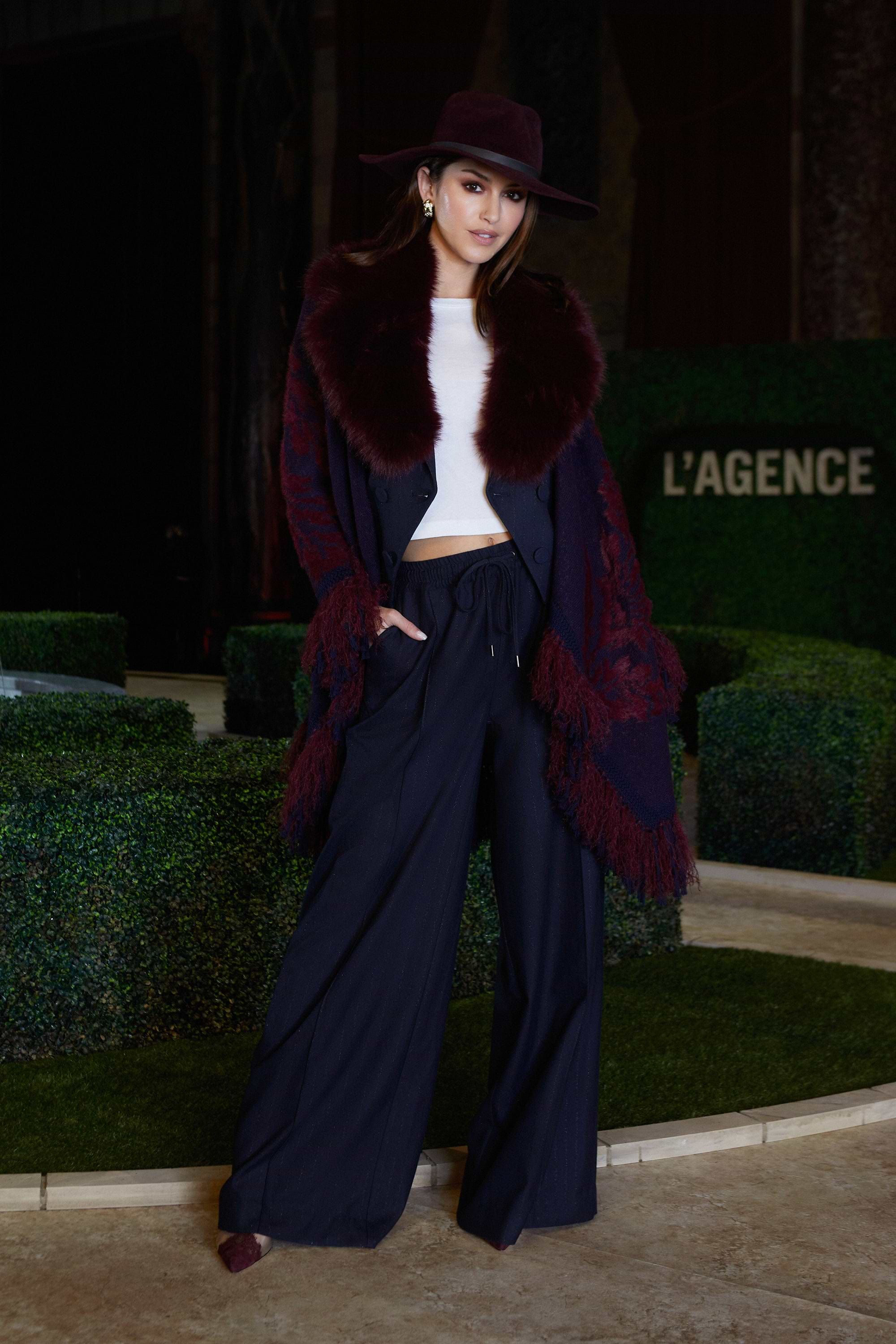 A woman poses confidently outdoors wearing a wide-brimmed hat, a white top, a dark fur-trimmed coat, and wide-leg navy pants. The background features manicured greenery and a LAGENCE sign.