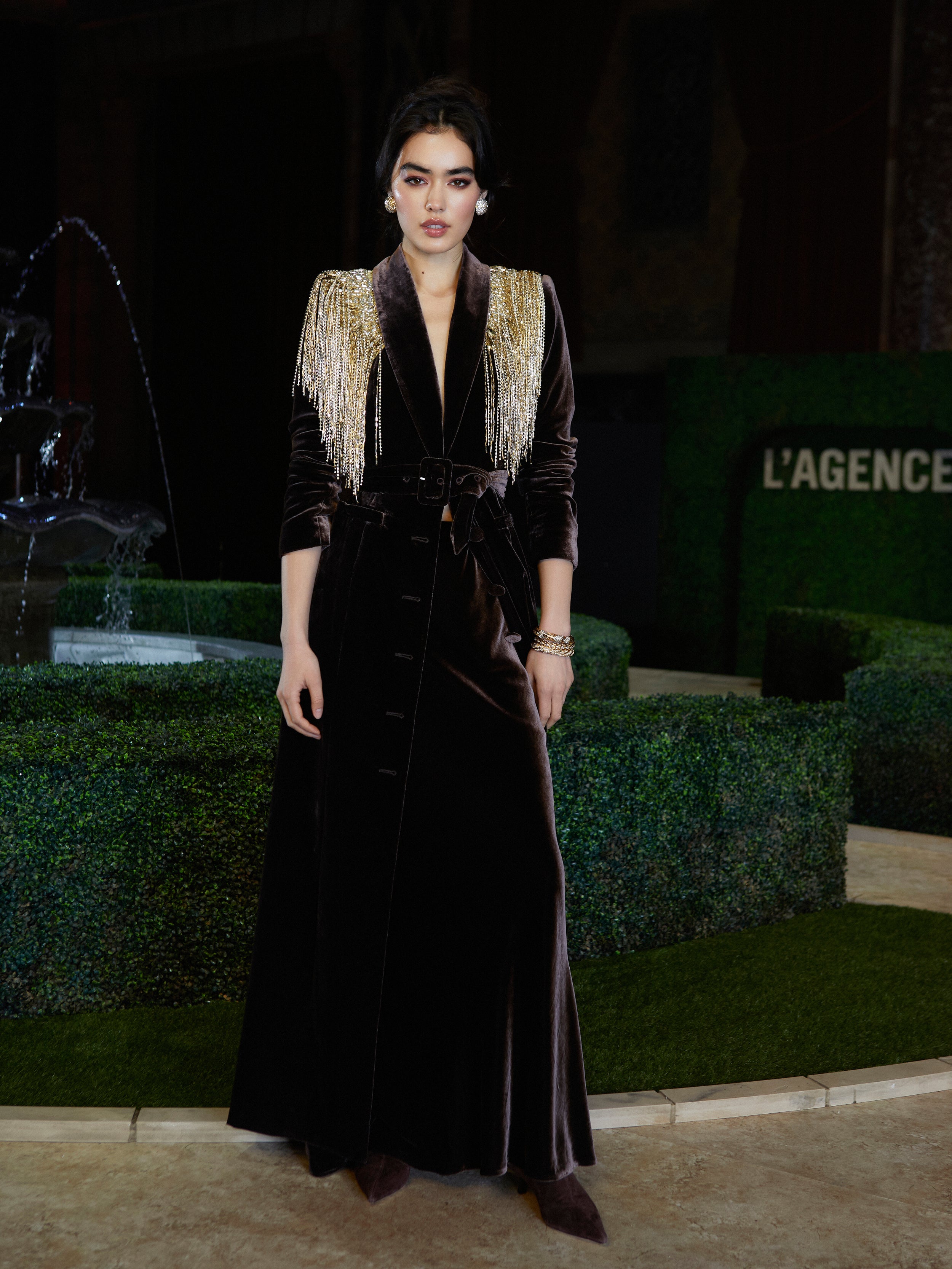 A woman wears the Raini Maxi Dress, a long dark velvet design with gold shoulder fringe, belt, and deep lapels, standing by sculpted greenery and a fountain at New York Fashion Week; behind her is an LAGENCE sign.