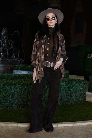 A stylish woman in wide-brimmed hat, sunglasses, and patterned jacket stands confidently by a decorative fountain. She wears a dark turtleneck, wide-leg pants, a statement belt, and gold jewelry.
