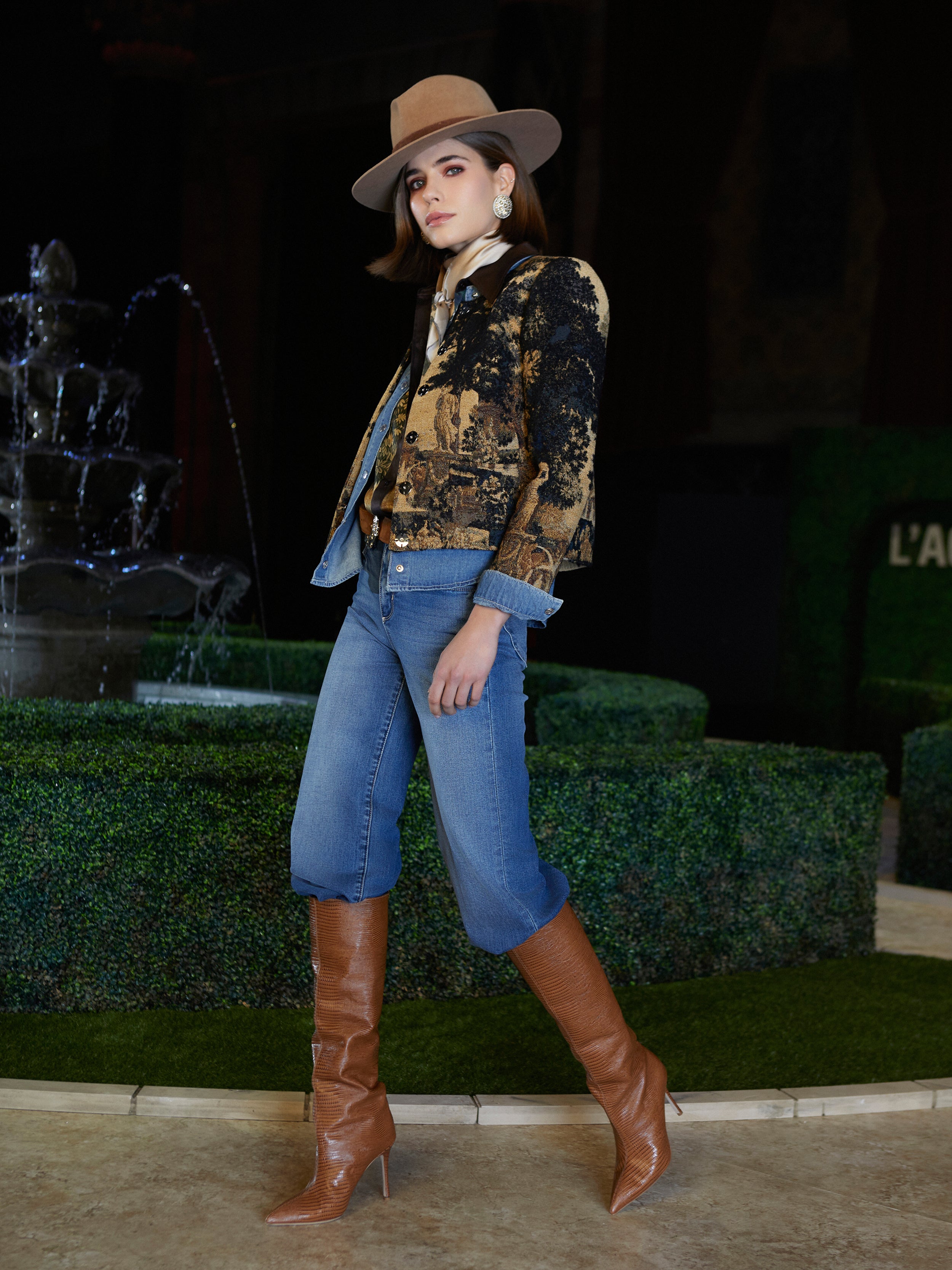 Channeling New York Fashion Week vibes, a woman poses by a fountain in the Isla Shirt, cuffed jeans, tall brown boots, and statement earrings. She exudes confidence against lush greenery—a vision for Fall 2026.