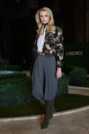 A woman wearing a sequined black jacket, white blouse, gray pleated harem pants, and knee-high boots stands in front of manicured hedges and a LAGENCE sign.