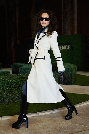 A woman with curly hair wears a long white coat with black trim, black gloves, black knee-high boots, and sunglasses. She strides confidently outdoors near manicured greenery, posing with one foot forward.
