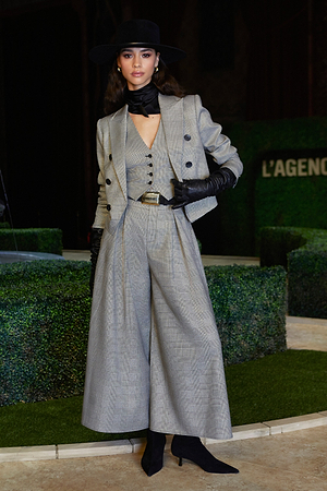 A woman wearing a stylish gray plaid suit with wide-leg trousers, a vest, and double-breasted blazer. She accessorizes with black gloves, a wide-brimmed hat, high-heeled boots, and a black scarf, posing confidently indoors.