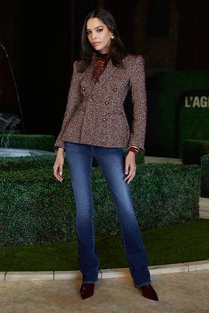 A woman stands confidently in a tailored maroon blazer, patterned blouse, blue jeans, and pointed burgundy shoes, with a fountain and manicured hedges in the background.