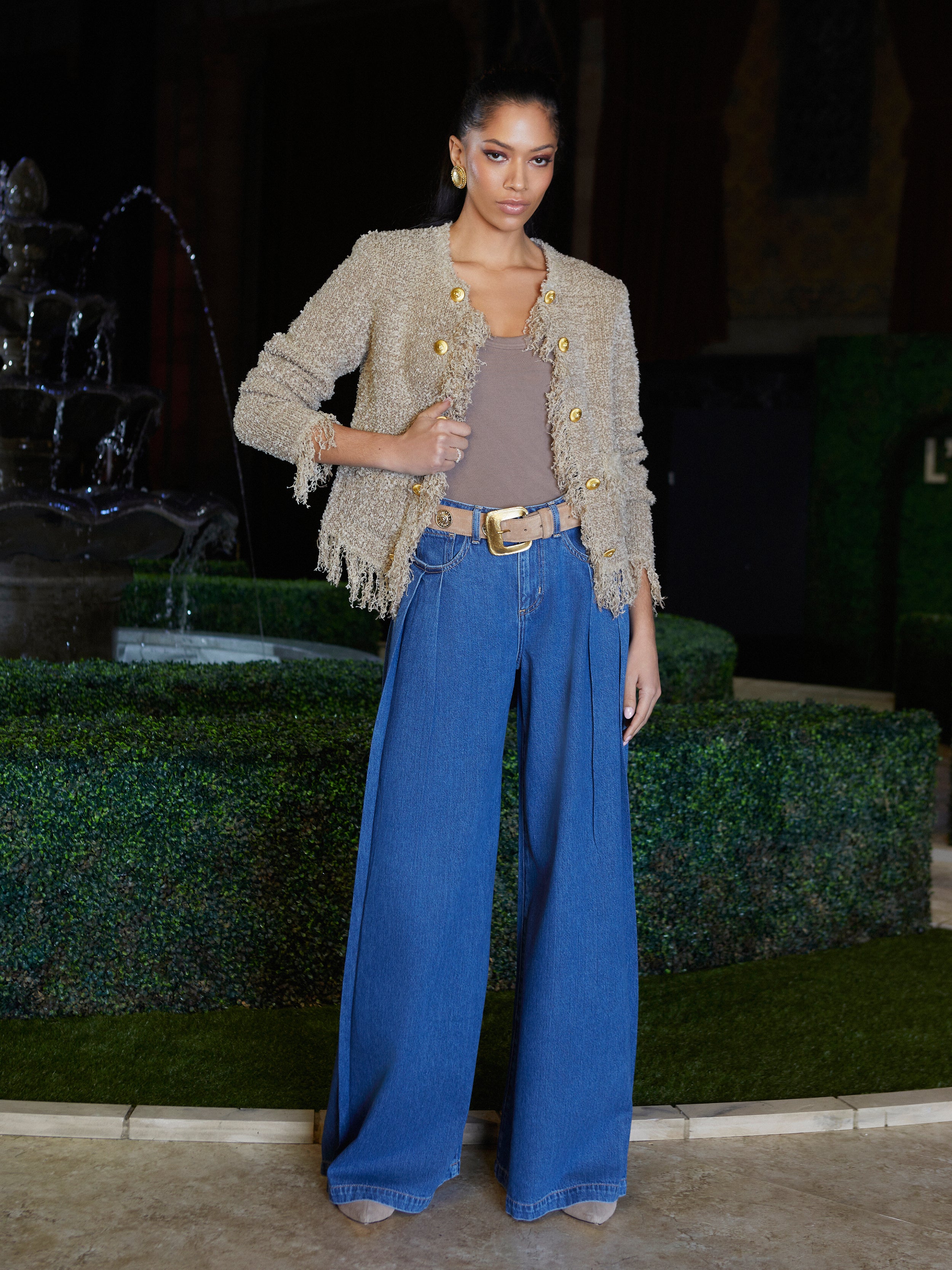 A woman poses confidently indoors wearing a textured beige jacket, a brown top, wide-leg blue jeans, and beige shoes. She stands in front of a trimmed green hedge and a decorative fountain.