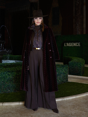 A woman in a dark brown wide-brim hat and Umbra Cabled Turtleneck stands by a fountain and manicured hedges at New York Fashion Week, showcasing the Fall 2026 fashion from L’AGENCE.