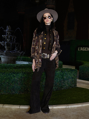 A woman exudes historical romance style in a wide-brim hat, sunglasses, a patterned jacket over a brown turtleneck, and the Livvy Suede Trouser as she stands indoors by a decorative fountain and manicured hedges.