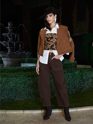 A woman stands confidently by a fountain in historical romance fashion, styled for Fall 2026 with heeled boots, a brown suede jacket, white shirt, corset, wide-brimmed hat, statement earrings, and Cody Cropped Barrel-Leg Jeans.