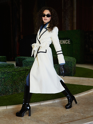 Wearing the Kamdyn Cropped Sweater, a woman with curly hair and sunglasses strides confidently along a tiled path bordered by manicured hedges, exuding Fall 2026 style.