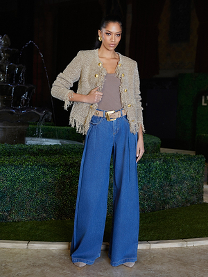 A woman poses indoors, radiating New York Fashion Week vibes in a textured beige jacket, the Shania Tank in brown, wide-leg blue jeans, and beige shoes, standing before a green hedge and fountain.