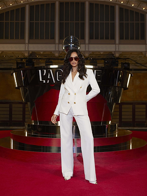 A woman in sunglasses stands confidently on a red carpet before a shiny black and silver LAGENCE backdrop, wearing the elegant Kyomi Double-Breasted Blazer. The glamorous indoor setting highlights her stylish outfit.