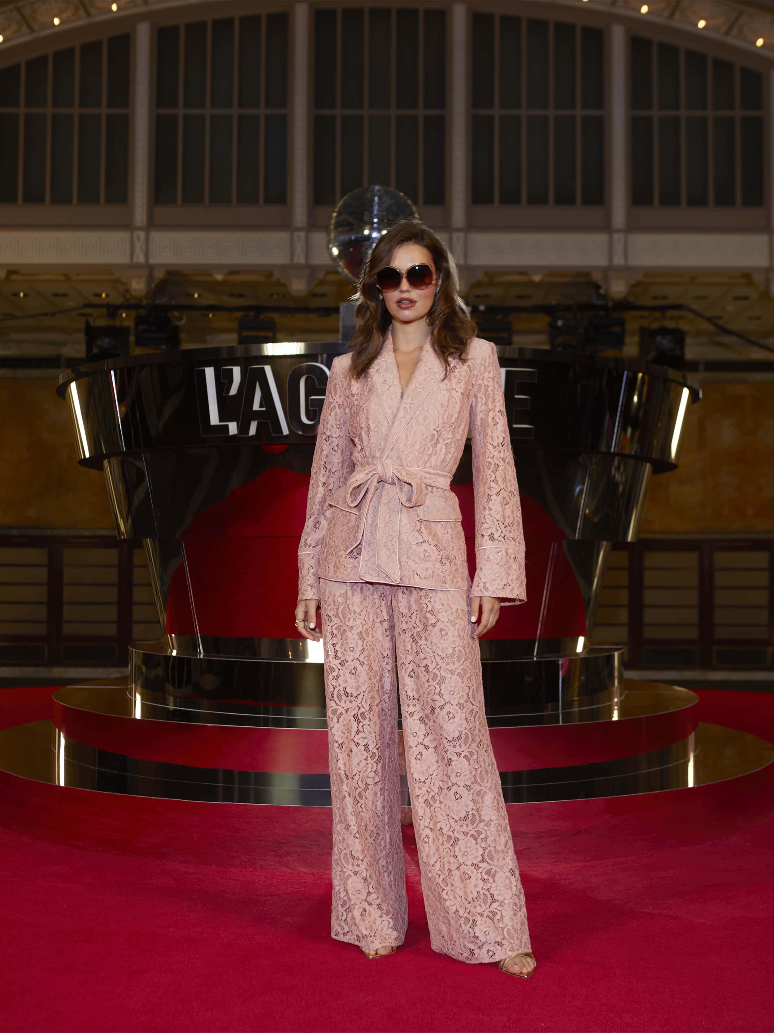 Wearing the Jordana Lace Blazer in light pink, a woman exudes confidence on the NYFW Spring 2026 red carpet before a mirrored L’AGENCE backdrop, channeling cinematic drama from their After Hours collection.