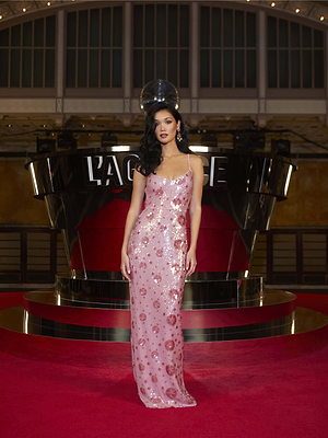 A woman in the Karma Sequin Maxi Dress, a shimmering pink gown, stands on a red carpet before a modern reflective backdrop with LAGENCE lit behind her. Her long dark hair and elegant posture are enhanced by the warm ambient lighting.