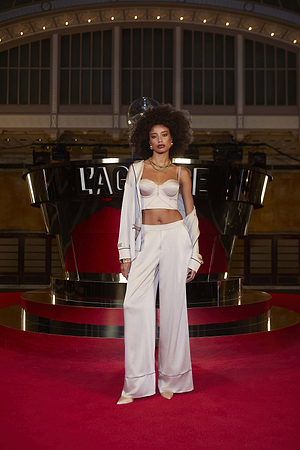 A woman with voluminous curly hair poses on a red carpet in the Mara Blouse, wide-leg trousers, and an open jacket before a shiny black and red “LAGENCE” backdrop.