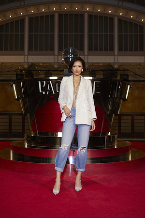 A woman stands confidently on a red carpet wearing a white blazer, layered necklaces, ripped blue jeans, and heels. Behind her is a shiny structure with LAGENCE displayed and a disco ball above.