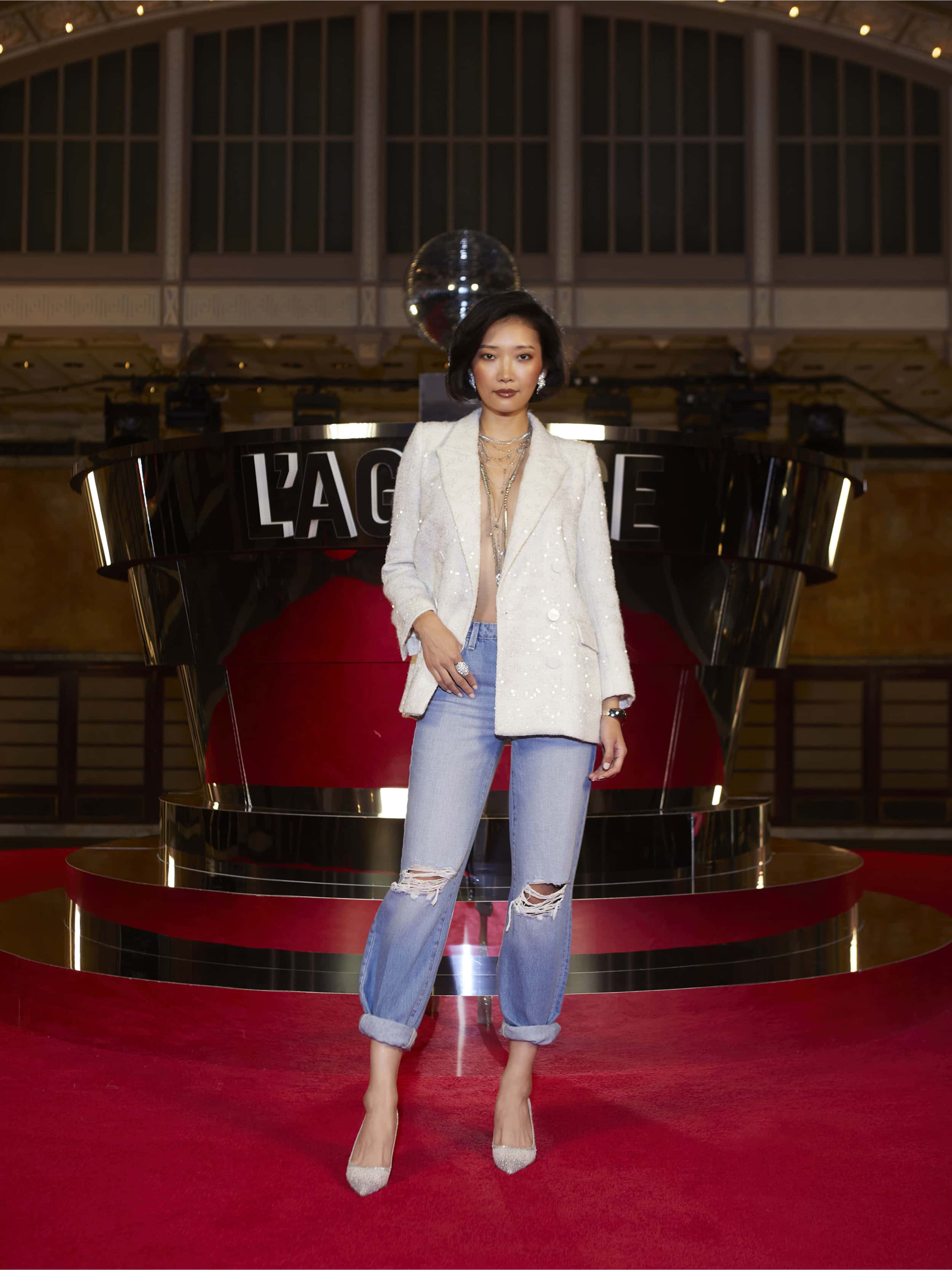 A woman poses on a red carpet in the Cambrie Oversized Sequin Blazer, layered necklaces, cuffed ripped jeans, and pointed heels—exuding cinematic drama at the LAGENCE Spring 2026 NYFW After Hours display.