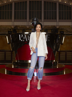A woman poses on a red carpet in a white blazer, layered necklaces, sheer top, and L'AGENCE Jones Stovepipe Jeans with ripped cuffs. The backdrop features a shiny black structure with L’AGENCE signage and a disco ball above.