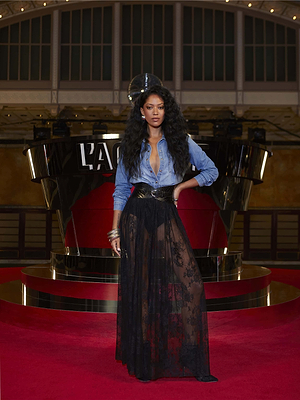 A woman with long curly hair poses on a red carpet in front of a modern LAMOUR display, wearing a denim shirt and the black Alona Lace Maxi Skirt with a wide belt.
