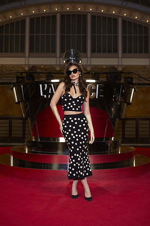 Wearing the Tula Knit Bra Top, she poses on the red carpet with wavy hair, black sunglasses, and heels, exuding cinematic drama against the shimmering LAVAZZA backdrop.