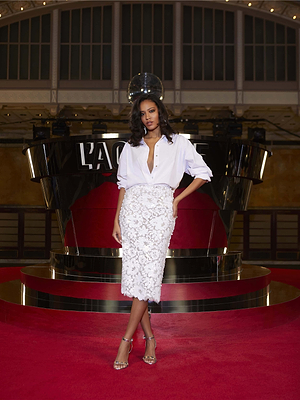 A woman stands on the red carpet in a crisp white button-up blouse, Makena Sequin Pencil Skirt, and silver heels. She poses confidently before a shiny black and gold backdrop—an elegant spring 2026 fashion statement.