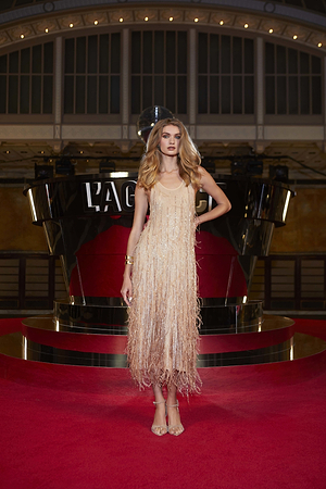 A woman in a beige, fringed flapper-style dress stands on a red carpet with one hand on her hip. She has long, wavy blonde hair and is posed in front of a shiny, black and gold art deco backdrop.