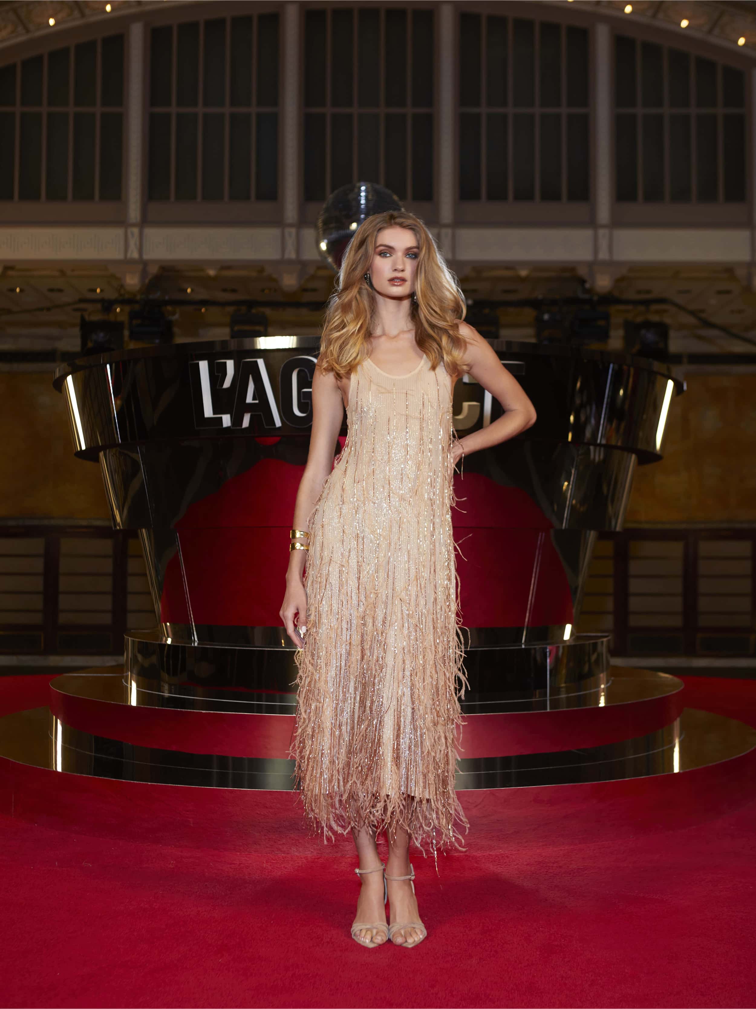 A woman in the Prince Dress and heels poses on a red carpet before a modern black and red art installation with LAGC above her, set against tall windows and ornate architecture.