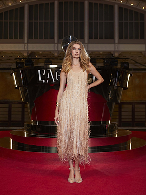 A woman in the Prince Dress and heels poses on a red carpet before a modern black and red art installation with LAGC above her, set against tall windows and ornate architecture.