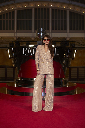 A woman wearing a sparkling beige pantsuit and sunglasses stands confidently on a red carpet, with her hand on her hip, in front of a modern, mirrored backdrop and a disco ball.