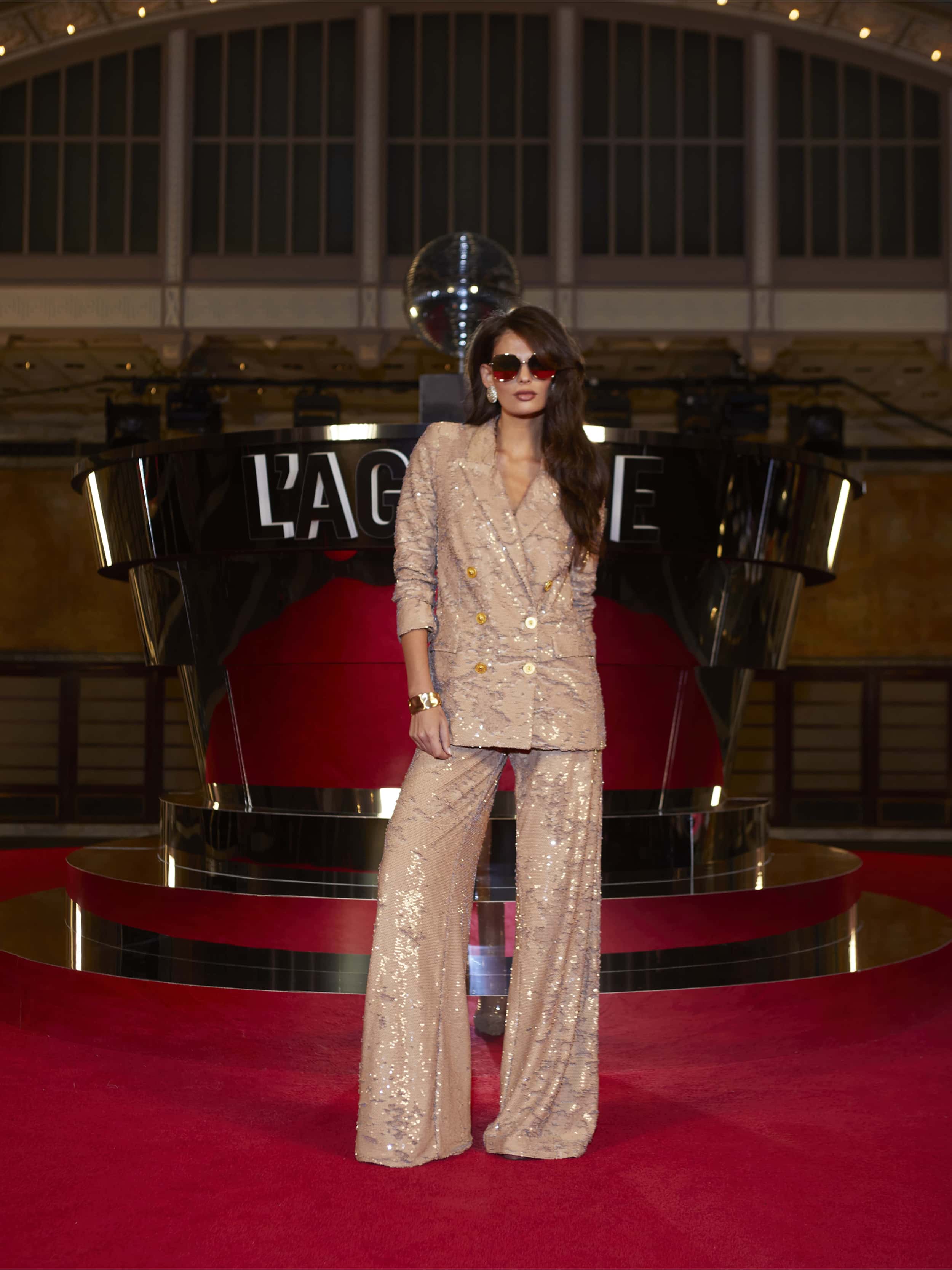 A woman in Tavia Lace Trouser and sunglasses stands confidently on a red carpet beneath marquee lights, channeling the bold spirit of After Hours at an NYFW show with a disco ball and metallic stage decor.