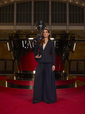 A woman wearing the Therese Blazer stands on a red carpet in front of a reflective black and chrome LAGENCE structure, inside an elegant venue with high ceilings and large windows.