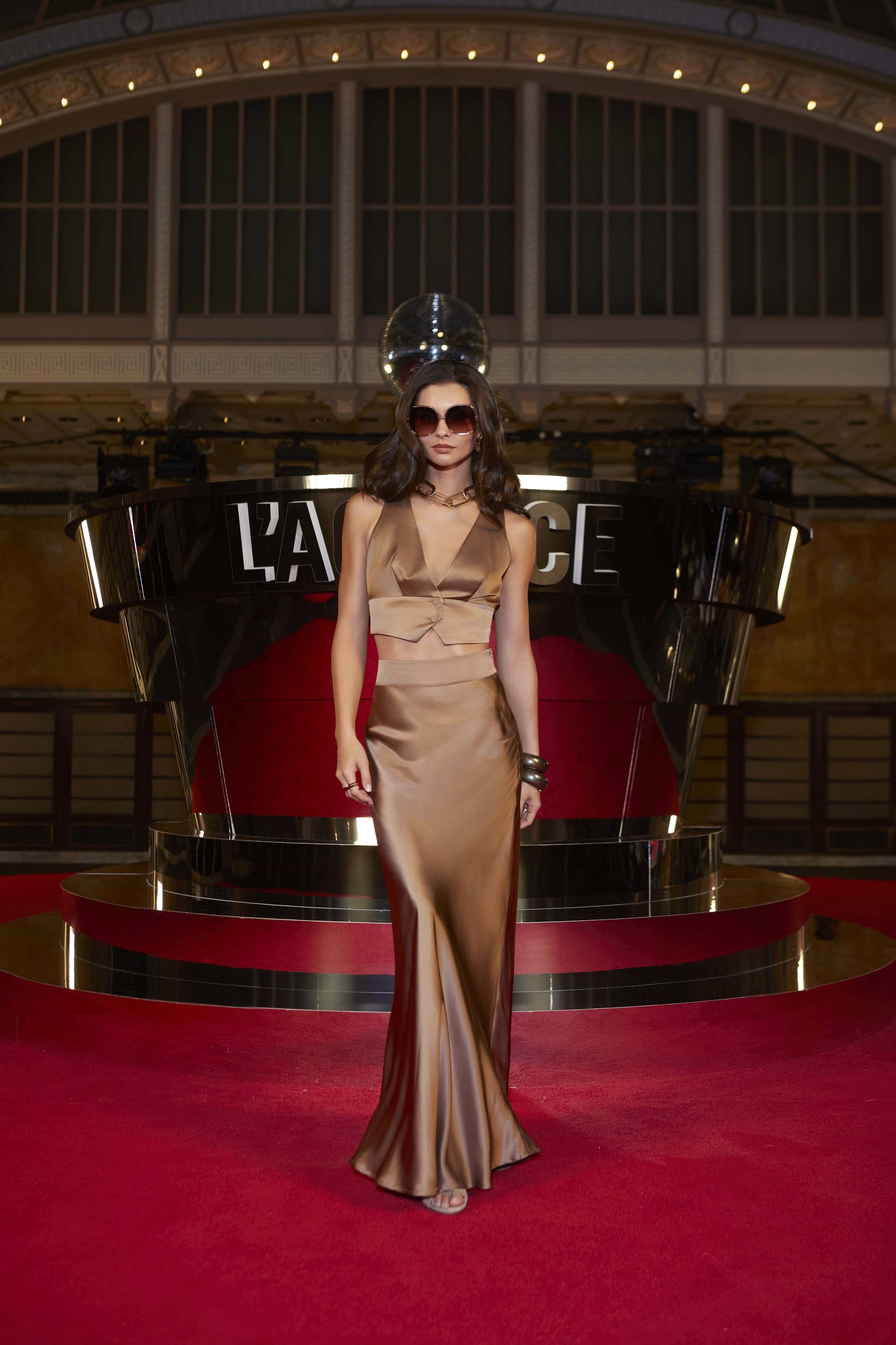 A woman in a gold satin gown and sunglasses stands on a red carpet in front of a building. A metallic structure with the word PALACE and a disco ball are behind her. The atmosphere is glamorous and dramatic.
