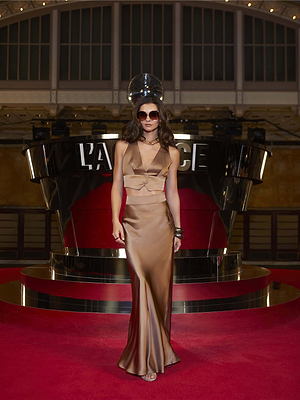 A woman in a shiny bronze evening gown and sunglasses poses on the red carpet before a black and chrome set with 