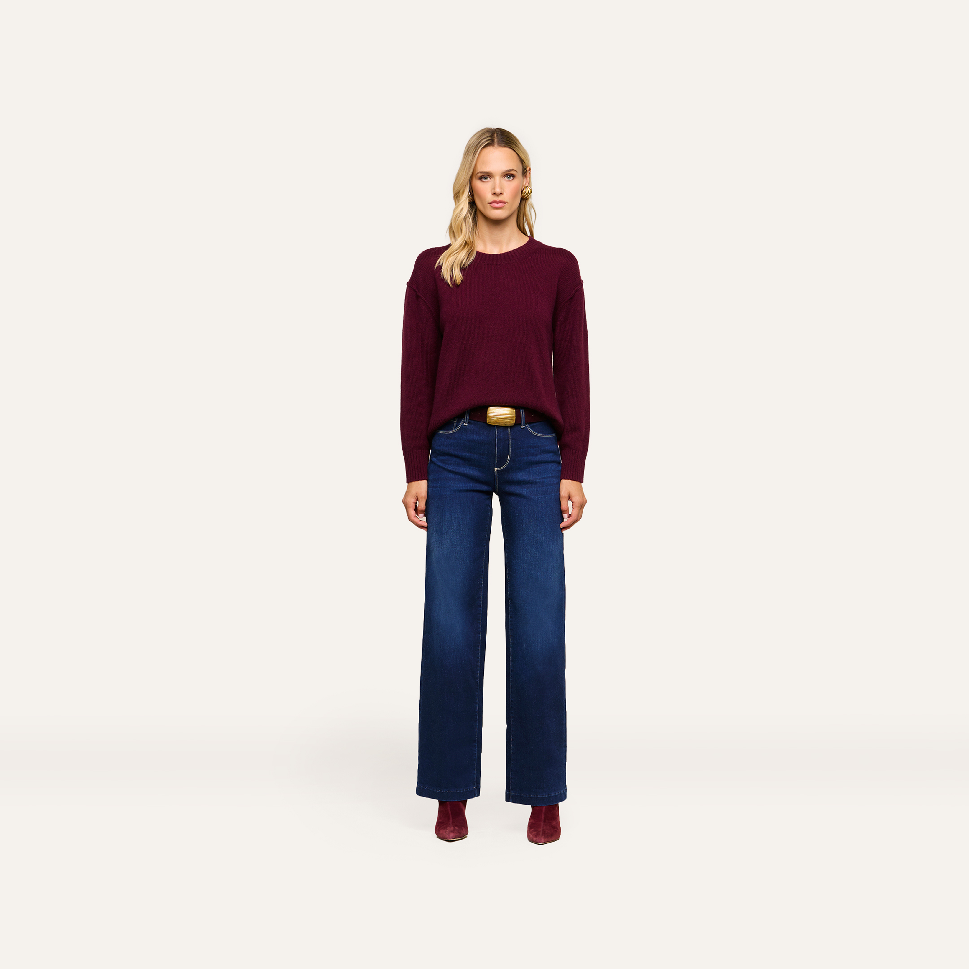 A woman stands facing forward, wearing a maroon sweater and wide-leg blue jeans with a belt, complemented by maroon boots, against a minimalist light background.