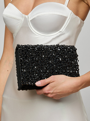 A person in a white satin dress holds the Victoire Crystal Clutch, a handcrafted rectangular black accessory adorned with shiny, textured beads—perfect for evening glamour and elevating any collection.