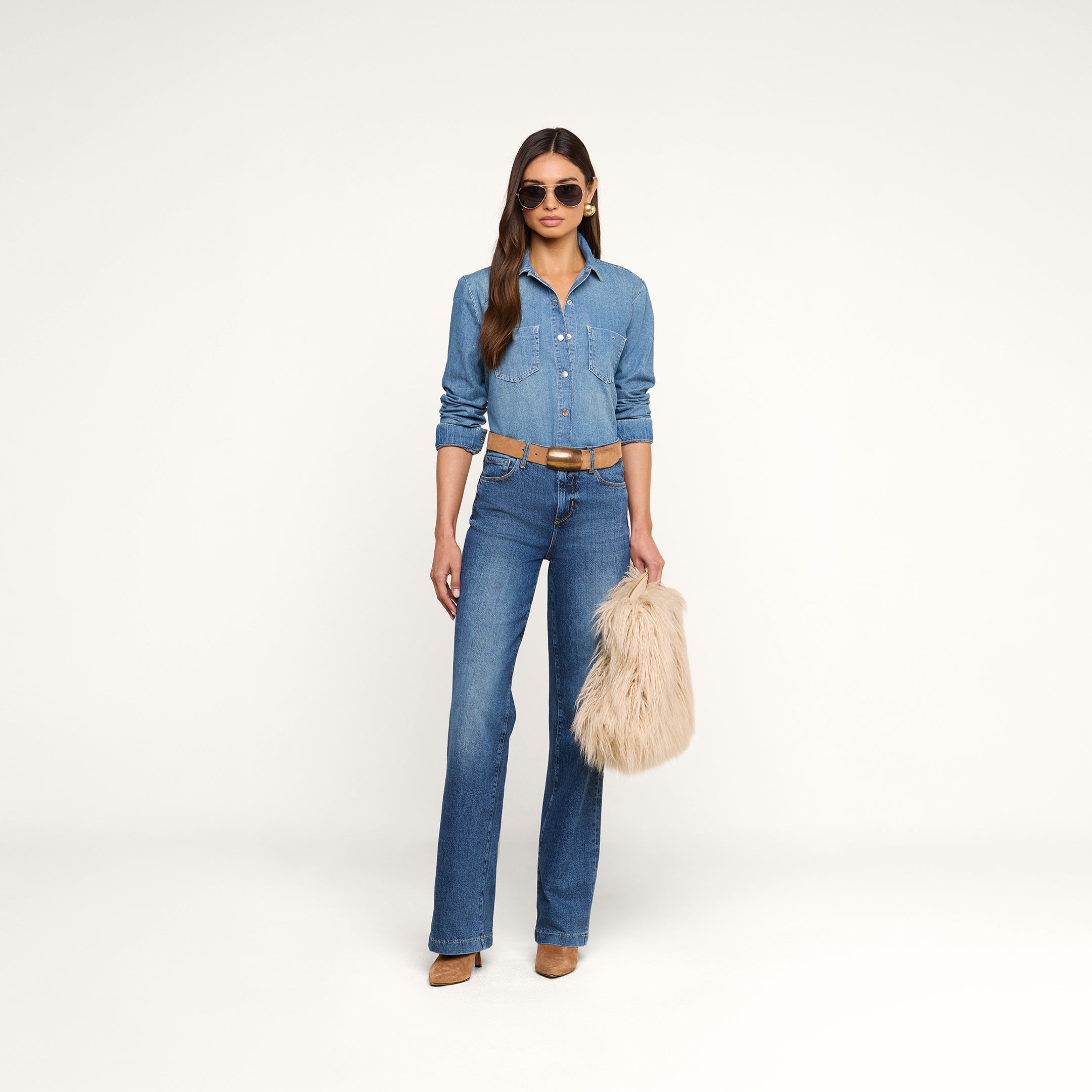 A woman with loose long hair stands against a white background wearing sunglasses, a denim shirt with jeans, brown heels, and a belt, holding the Adelina Faux Fur Clutch.