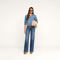 A woman stands against a white background wearing a denim shirt, blue jeans, tan belt, tan boots, and sunglasses, holding the Adelina Faux Fur Clutch under her arm.
