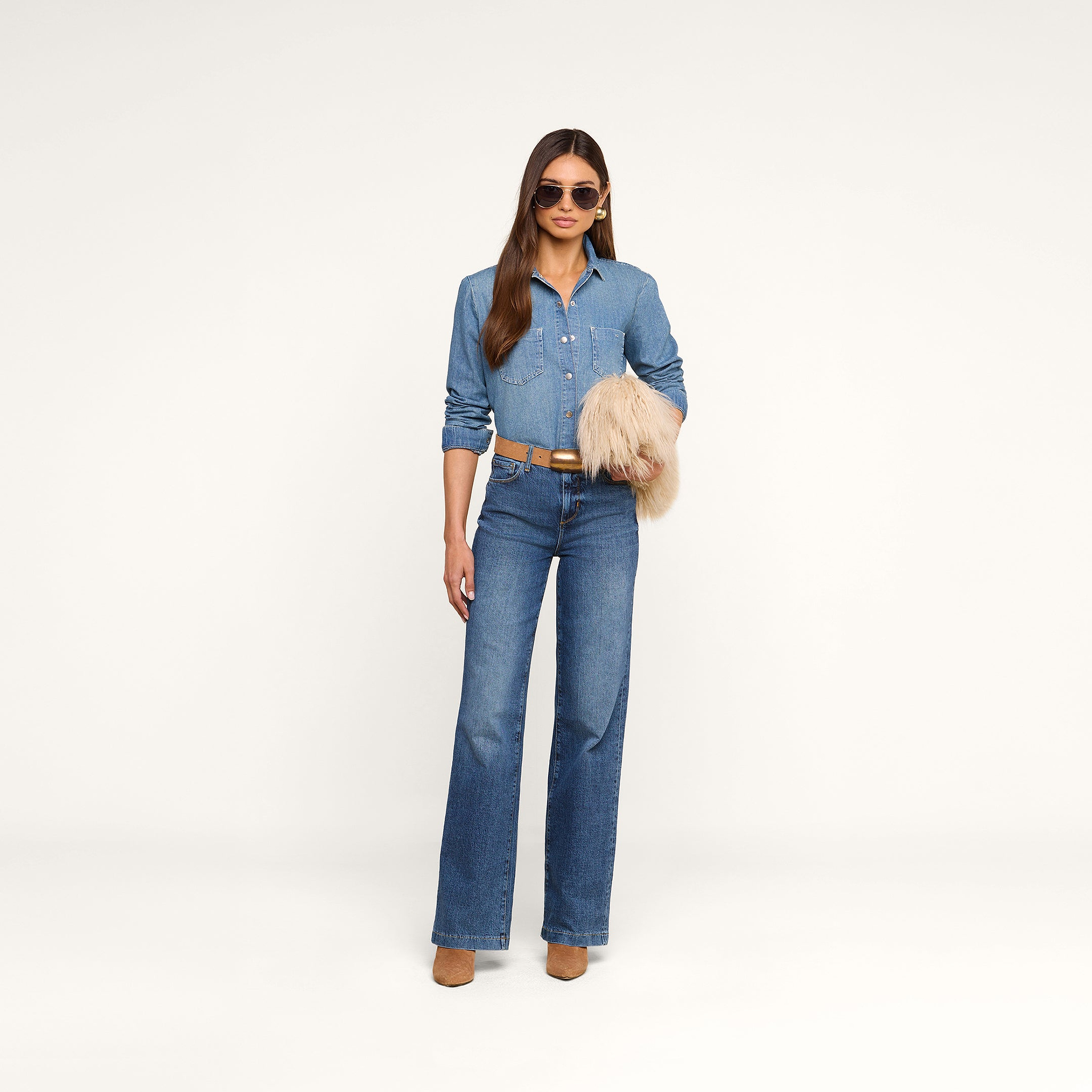 A woman stands against a white background wearing a denim shirt, blue jeans, tan belt, tan boots, and sunglasses, holding the Adelina Faux Fur Clutch under her arm.