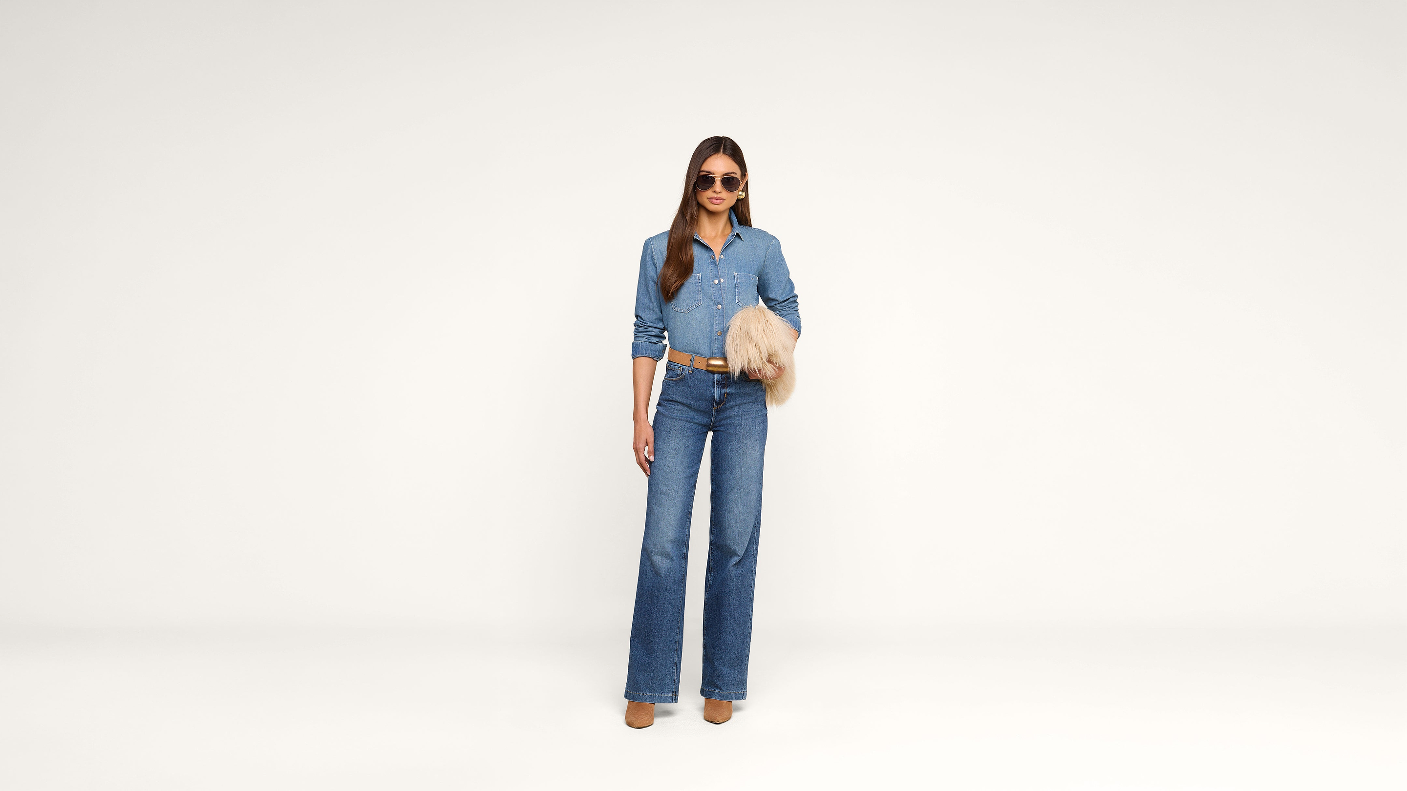 A woman stands against a white background, wearing sunglasses, a blue button-up shirt, wide-leg jeans, tan boots, and holding a tan fur accessory.
