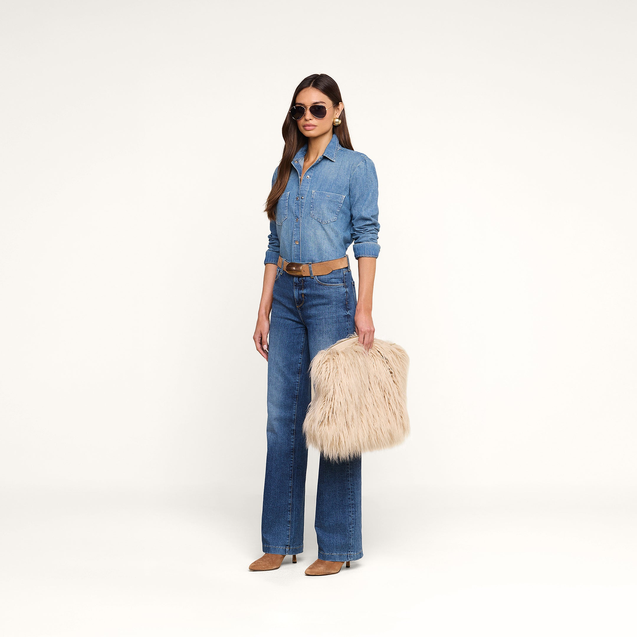 A woman in a denim shirt and jeans, wearing sunglasses and brown shoes, stands holding the Adelina Faux Fur Clutch against a plain white background.