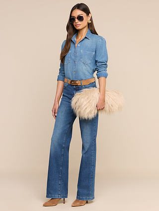 A woman stands against a plain beige background, dressed in a denim shirt and jeans paired with a tan belt and tan pointed-toe shoes. She accessorizes her look with aviator sunglasses and carries the Adelina Faux Fur Clutch in beige as a statement piece.