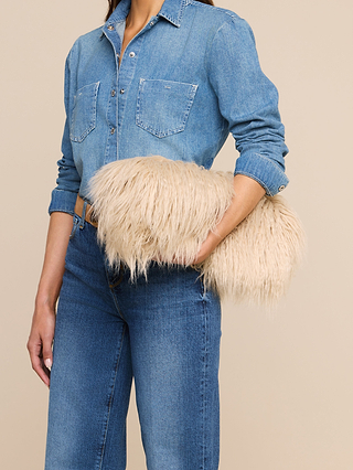 A woman dressed in a denim shirt and jeans holds the Adelina Faux Fur Clutch—a large, fluffy beige statement accessory—under her arm against a plain tan background.