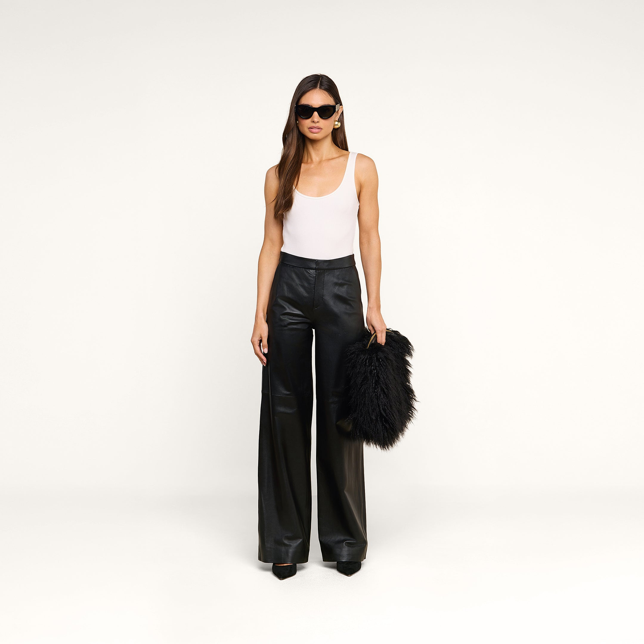 A woman stands against a plain background, wearing sunglasses, a white tank top, and black wide-leg pants while holding the Adelina Faux Fur Clutch in her left hand.