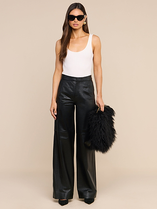 A woman stands against a beige background, wearing sunglasses, a white tank top, black wide-leg pants, and black heels while holding the Adelina Faux Fur Clutch as a statement accessory.
