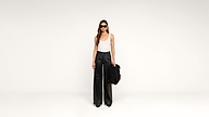 Woman in sunglasses, a white tank top, and black wide-leg pants stands against a white background, holding a large black furry bag by her side.