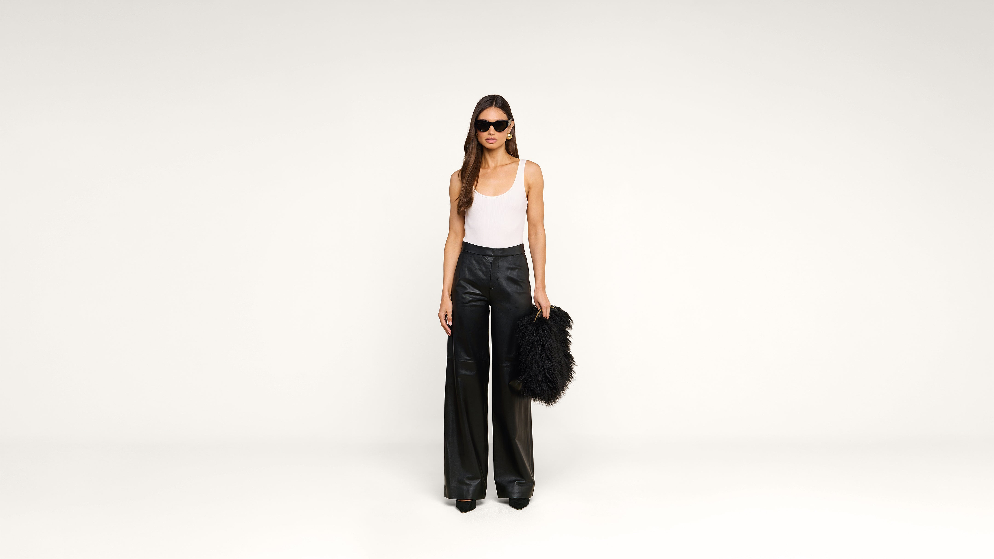 Woman in sunglasses, a white tank top, and black wide-leg pants stands against a white background, holding a large black furry bag by her side.