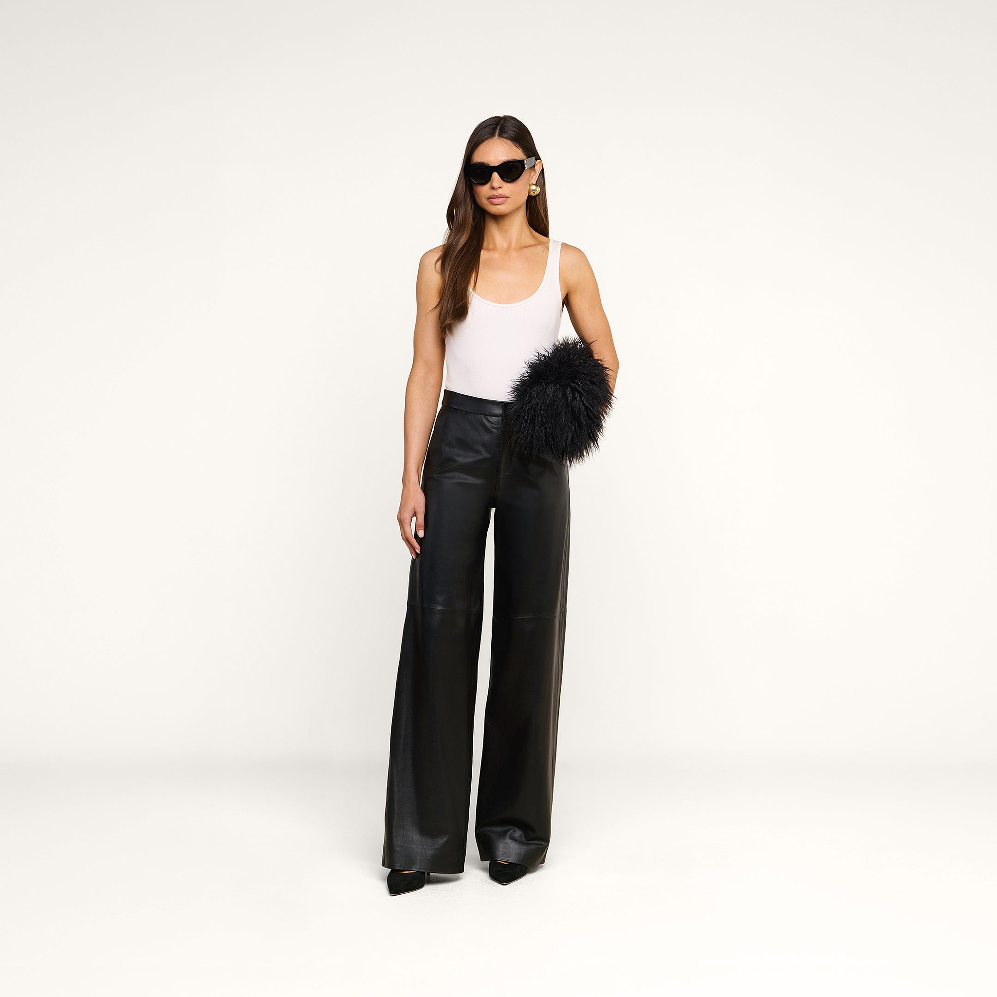 A woman in sunglasses wears a white tank top, black wide-leg pants, black heels, and holds the Adelina Faux Fur Clutch against a plain white background.