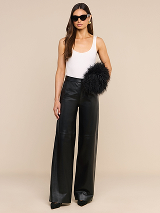 A woman wearing sunglasses, a white sleeveless top, high-waisted black leather wide-leg pants, and holding the Adelina Faux Fur Clutch as a statement accessory stands against a plain beige background.